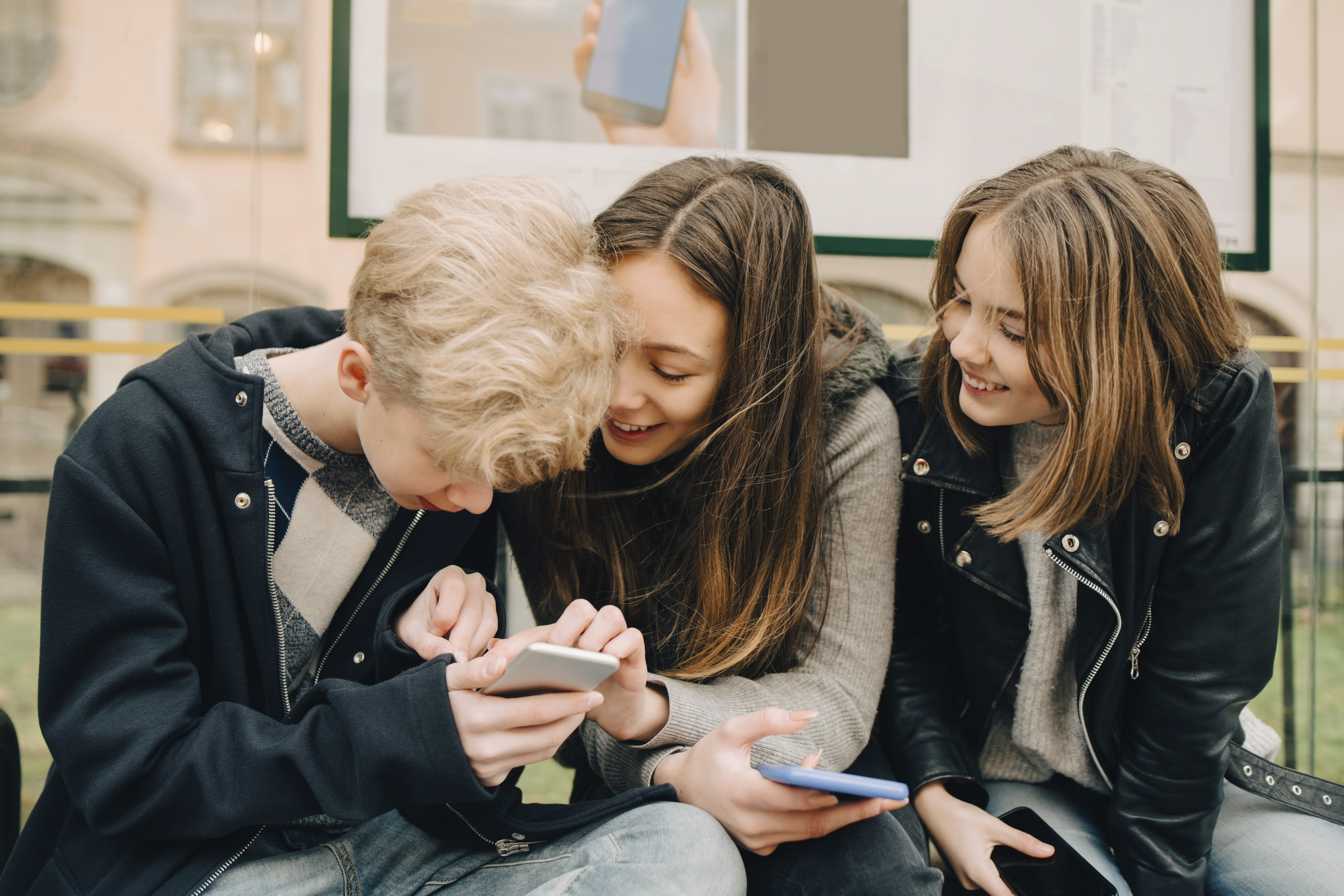 En grupp leende ungdomar sitter på en busstation och tittar tillsammans på en mobiltelefon